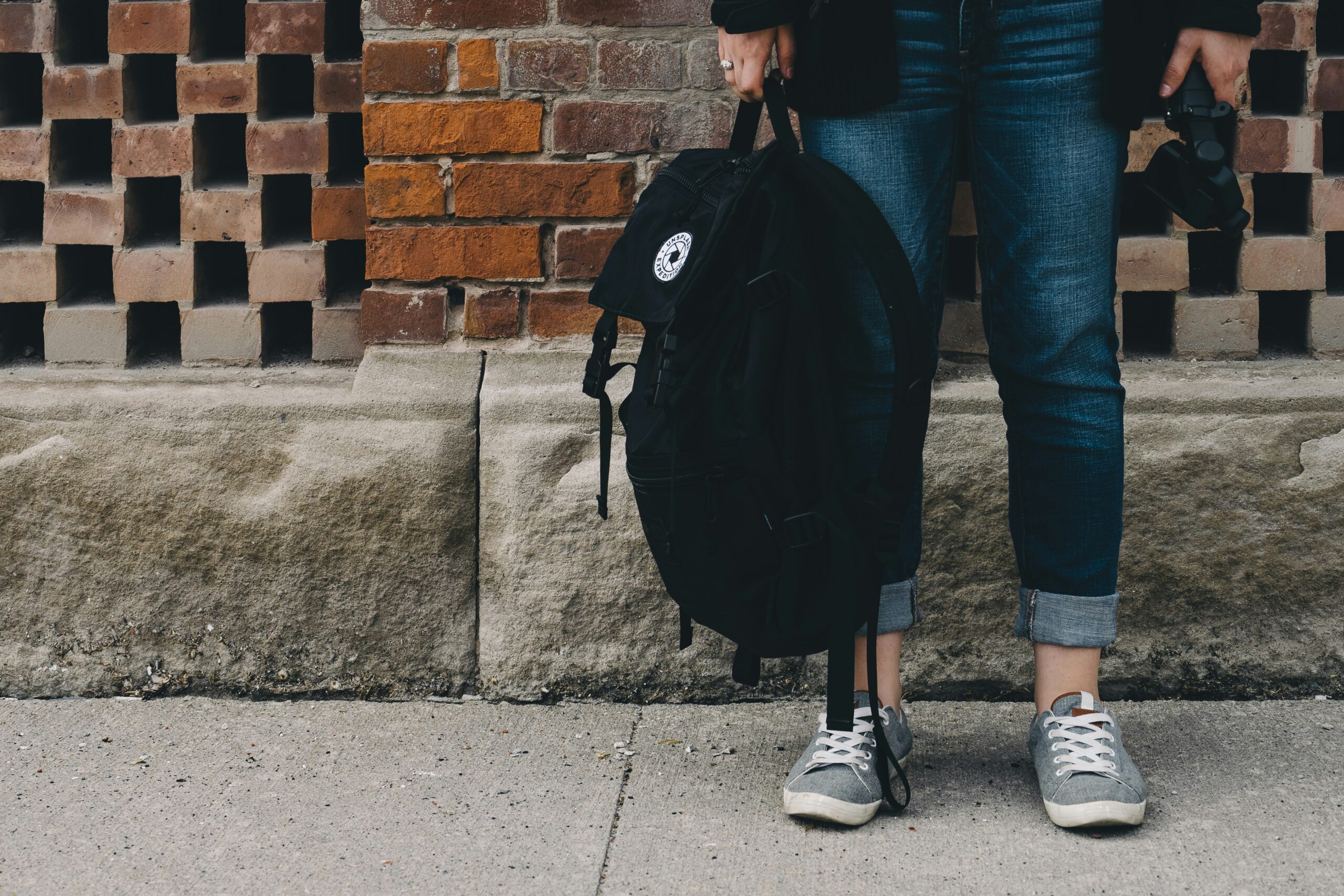 woman standing you can only see front her waist down on the street sidewalk wearing jeans holding a black backpack in front of a brick wall