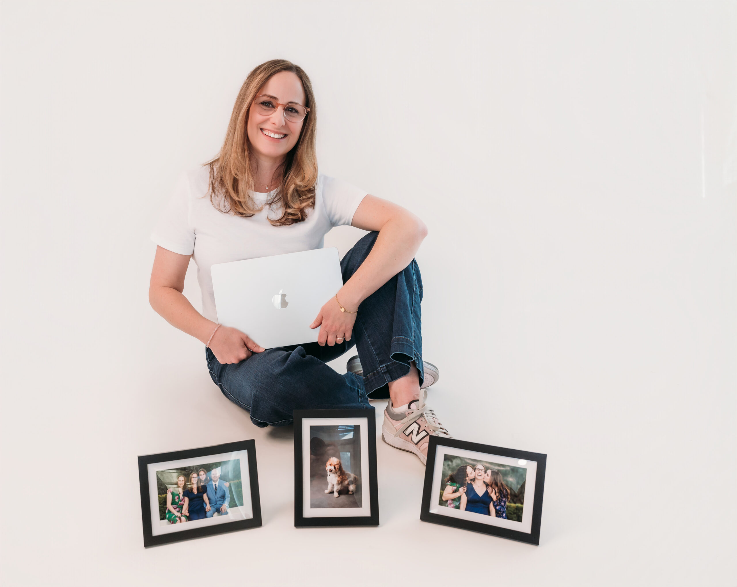 A woman in a white shirt and jeans sits on the floor holding a laptop, with three framed photos placed in front of her against a plain white background.
